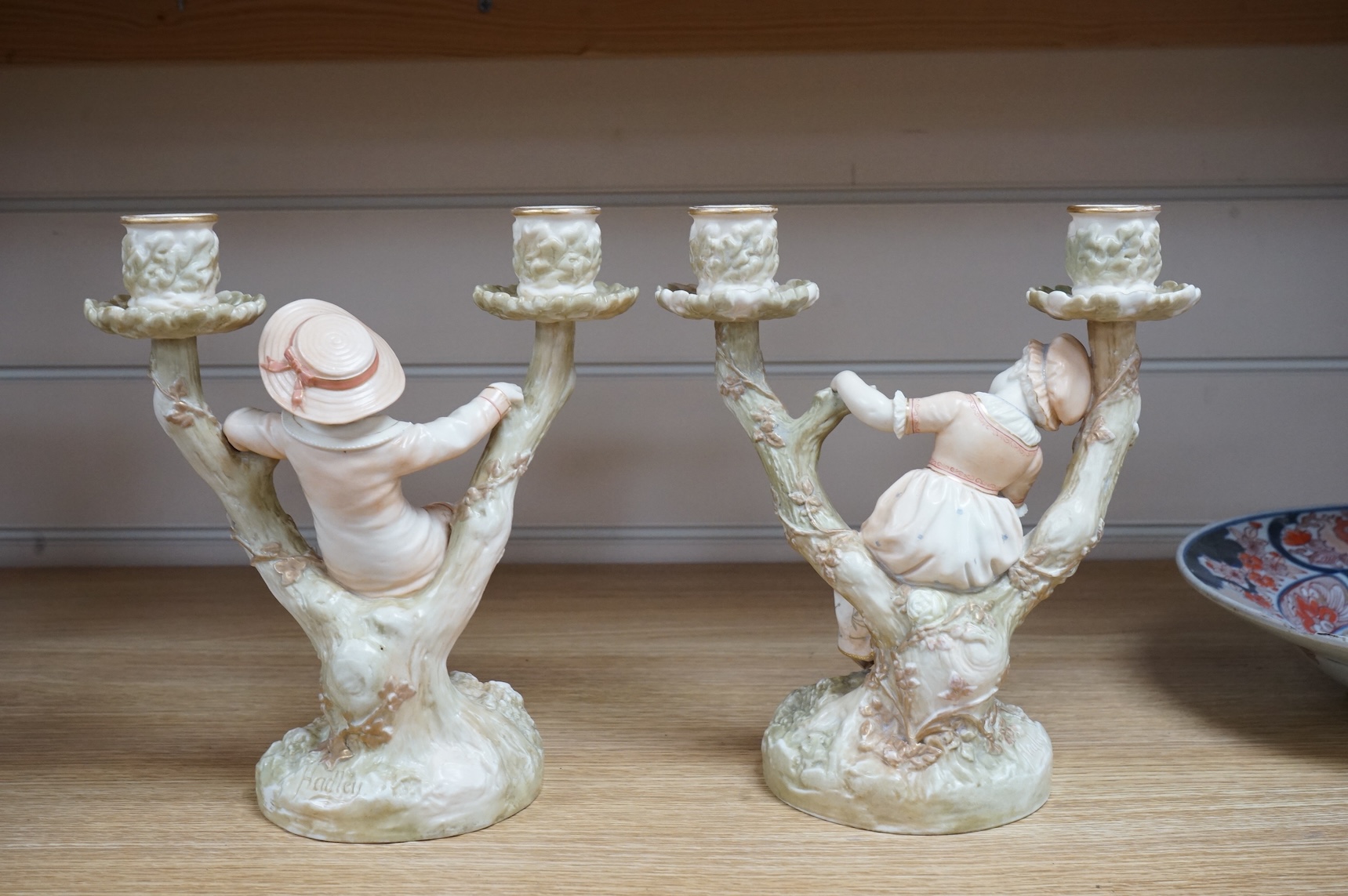 A pair of late 19th century Royal Worcester Kate Greenaway style figural two branch candelabra, modelled by James Hadley 22.5cm high